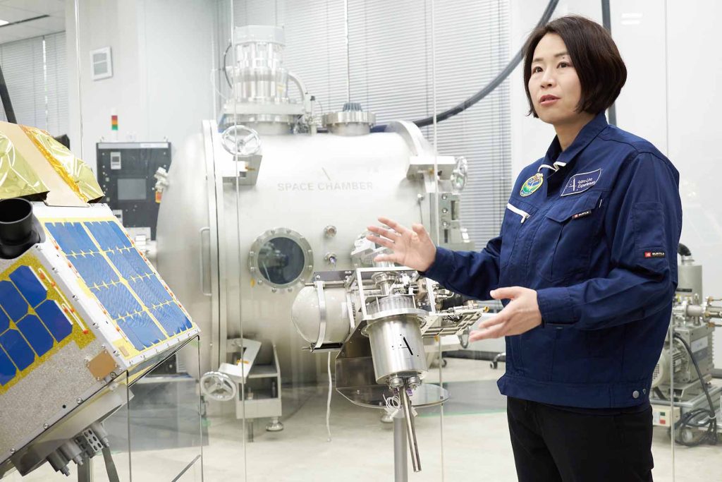 人工衛星（左）と流星源を射出するユニット（中央）の模型を説明する岡島さん。奥の実験用クリーンルーム内にあるのは、真空を再現する装置